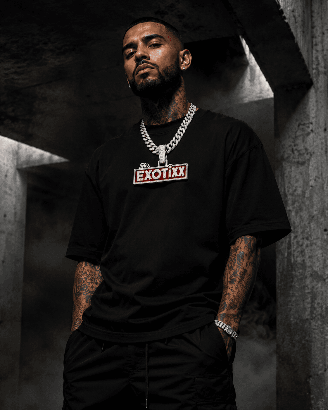 Model in a black Mr. Exotixx logo-box tee with silver chains in a brutalist concrete environment