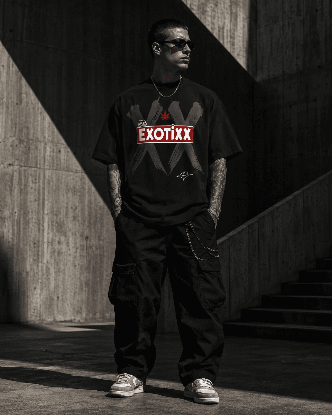 Model in black Mr. Exotixx XX-graphic tee and cargo pants standing in a sunlit concrete corridor