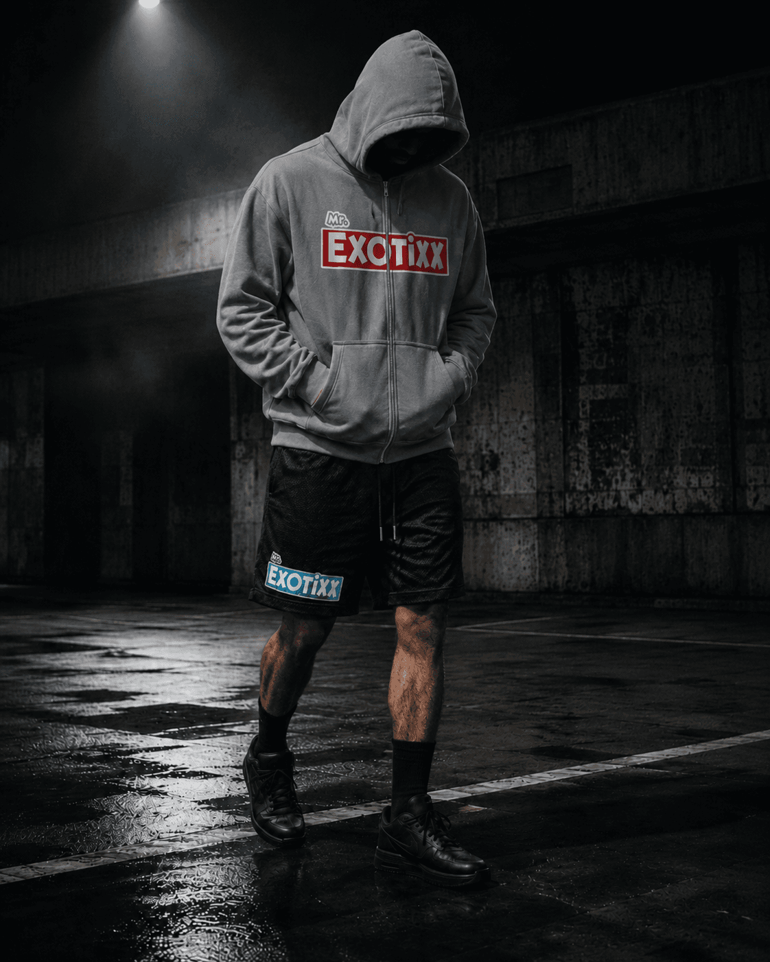 Figure in gray Mr. Exotixx zip-up hoodie and branded shorts under a single overhead light